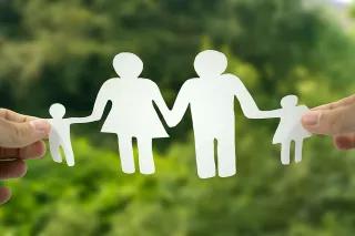 Dies ist ein Foto eines Scherenschnitts in Form einer Familie (zwei Eltern, zwei Kinder), der vor einen grünen Waldhintergrund gehalten wird.